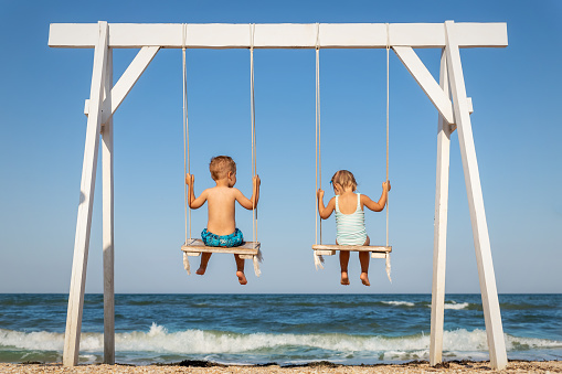 Back view couple of cute little adorable small caucasian sibling kids enjoy having fun swinging and playing at playground sea sand beach against blue ocean water wave and clear sky. Family vacation.