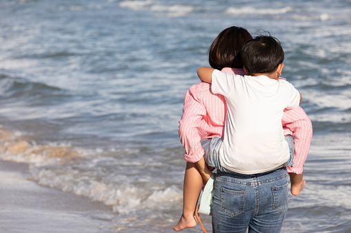 back view mother carrying her son on back and walking on the beach