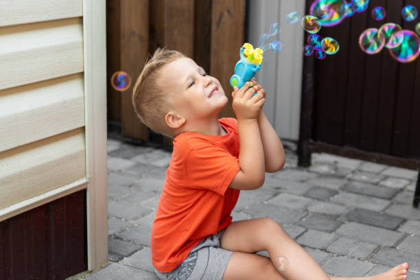 porträt eines süßen kleinen bond-kind-jungen, der spaß daran hat, seifenblasen zu spielen, die an einem hellen, warmen sommertag an der hauswand im freien sitzen. kinder gesund außerhalb der natur aktivitäten - child bubble wand children only park stock-fotos und bilder