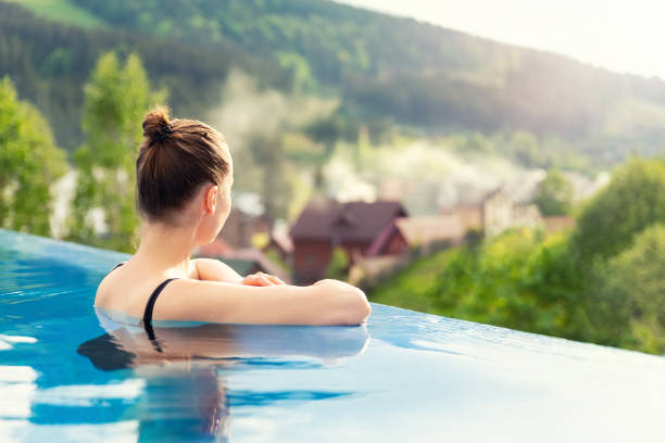 back view young adult female person enjoy relax in infinity edge luxury outdoor swimming pool looking on fog hill green mountains in warm sunset light. Alpine welness travel vacation tourism concept stock photo