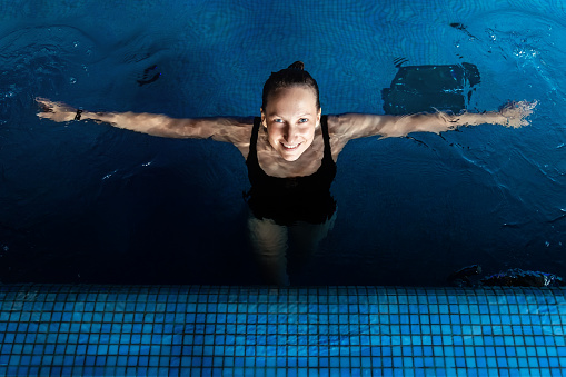 Above top view portrait of young adult caucasian female model in sport black swimsuit stand in clear blue water of indoor swimming pool at dark evening time. Relax harmony and wellness spa therapy.