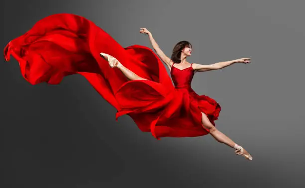 Photo of Ballerina Dance. Ballet Dancer in Red Dress jumping Split. Woman in Ballerina Shoes dancing in Silk Gown flying on Wind over Gray Studio Background