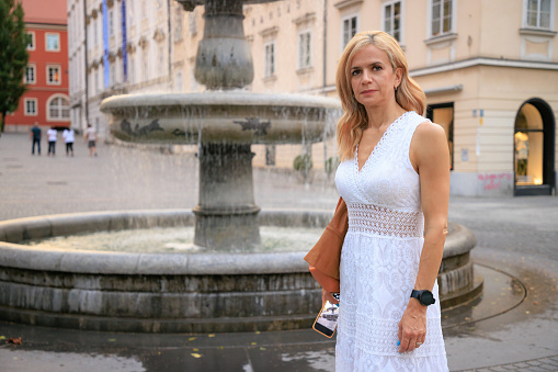Woman in white summer dress with phone in hand standing in front of the fountain in the historical part of the city, city break in Ljubljana