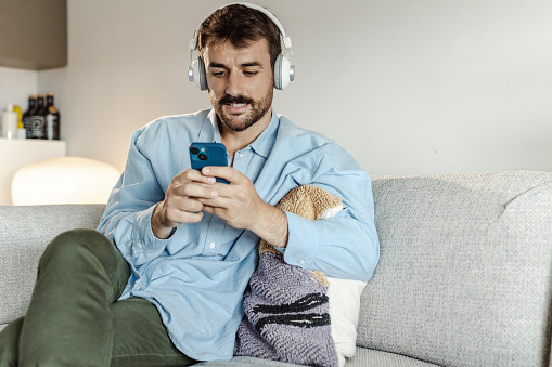 Portrait of a bearded man concentrating on scrolling social media feed. He is sitting on the couch in the living room and listening music or podcast via wireless headphones.