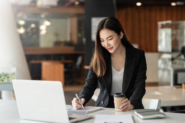 istock Charming Asian businesswoman holding a coffee cup and taking notes on laptop and document at office. 1404178485