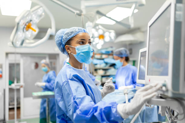 anesthetist working in operating theatre wearing protecive gear checking monitors while sedating patient before surgical procedure in hospital - ziekenhuis stockfoto's en -beelden