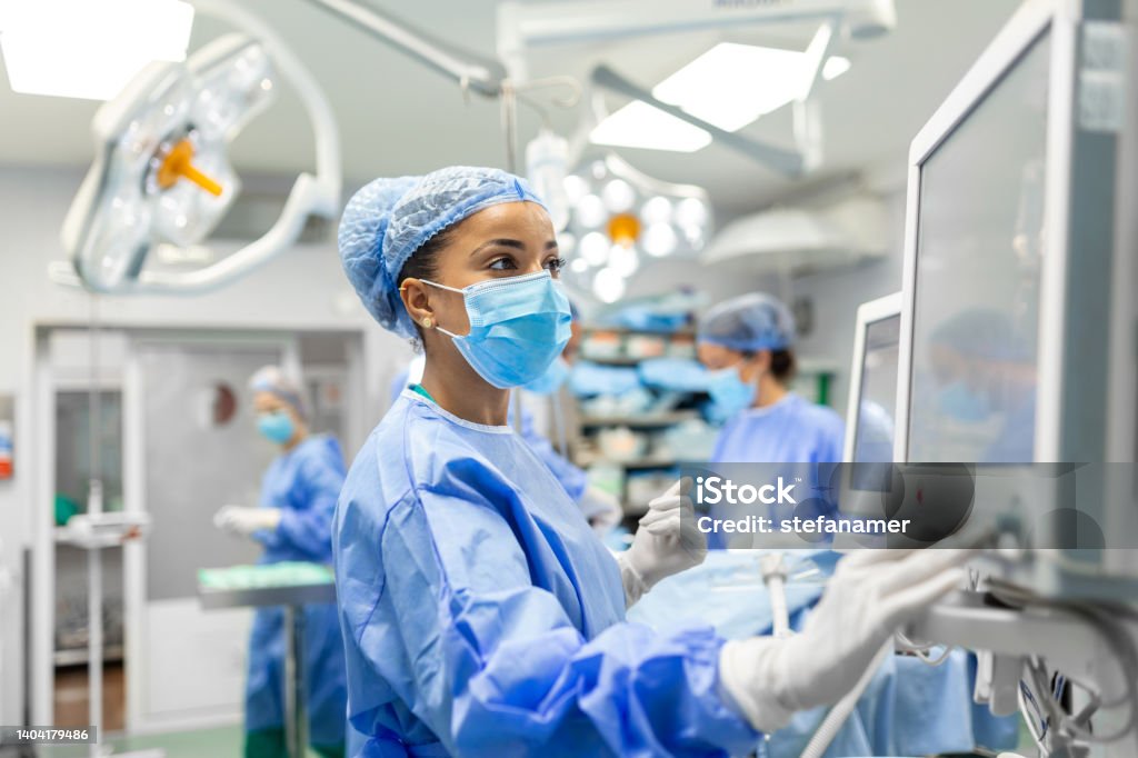 Anesthetist Working In Operating Theatre Wearing Protecive Gear checking monitors while sedating patient before surgical procedure in hospital Nurse Stock Photo