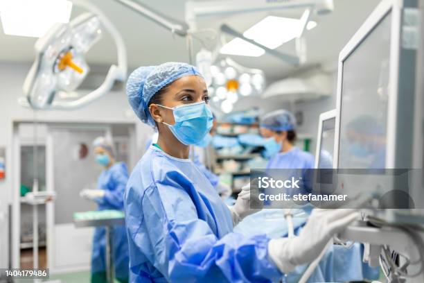 Anesthetist Working In Operating Theatre Wearing Protecive Gear Checking Monitors While Sedating Patient Before Surgical Procedure In Hospital Stock Photo - Download Image Now