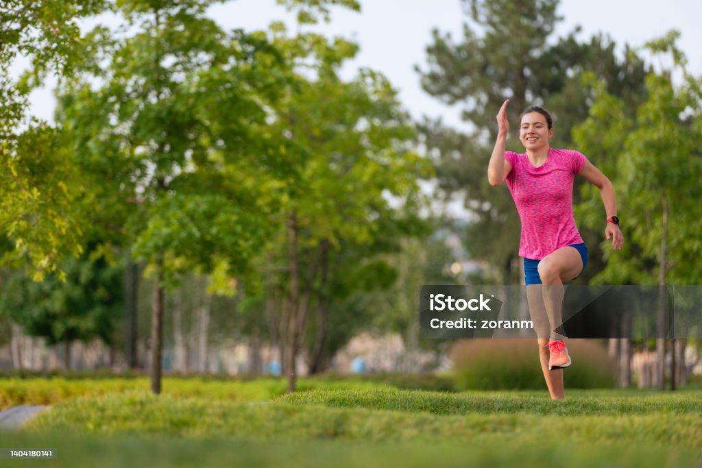 Smiling young woman in running shorts jogging on grass padded track in public park Happy young woman wearing running shorts jogging on grass padded track in public park in the city, healthy lifestyle in motion 18-19 Years Stock Photo