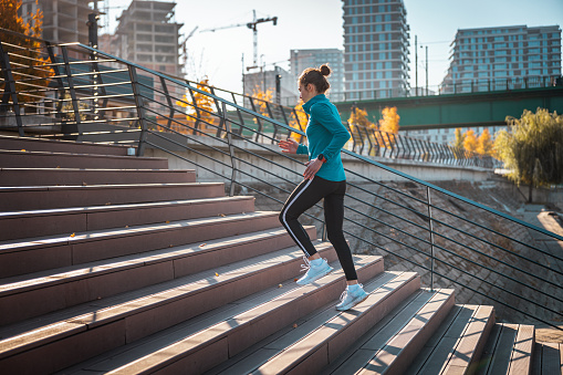 Young woman in sports clothes running up the stairs on the city waterfront, residential buildings construction site in the background, healthy lifestyle in the city