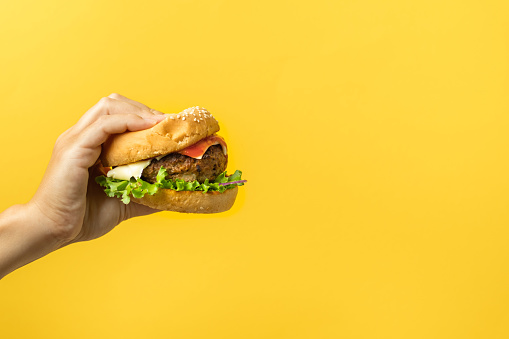 Woman hand holding a burger on a yellow background. Free space for text. Dieting concept