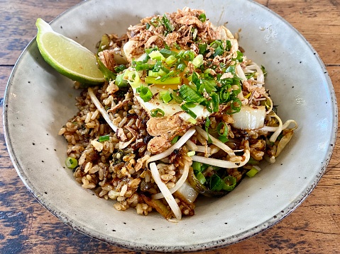 Horizontal close up of handmade ceramic bowl of organic nasi goreng fried rice with fresh herbs, whole fried egg, lime paste and seeded sprouts on wood table