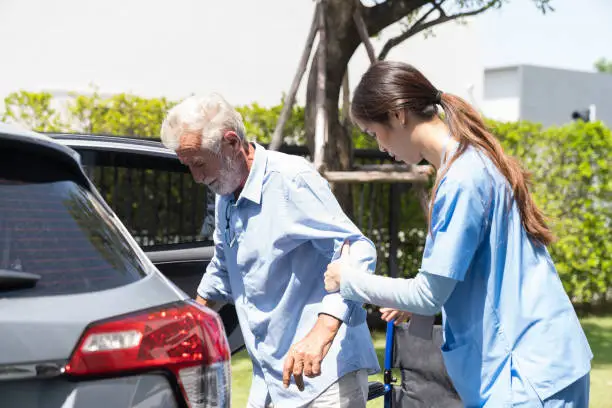 Female nurse taking care careful senior man on wheelchair into car. Caring Asian nurse taking care of elderly man in wheelchair at the hospital ward outdoor. People and health care concept