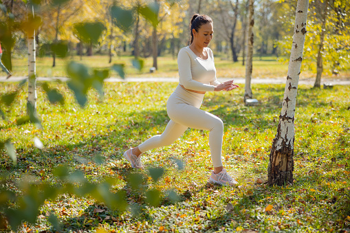 Asian woman in casual sports clothes assuming lunge position for stretching in the park, leisure outdoor workout on sunny autumn day
