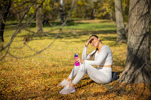 Asian woman in casual sports clothes sitting on dry leaves in the forest by the tree, smiling and shielding eyes from sunlight with hand, relaxing after workout outdoors