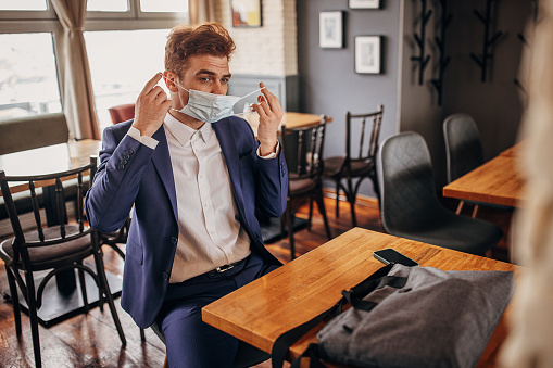Handsome young businessman sitting in cafe he is putting on a protective face mask.