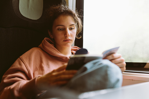 teenage girl in sweatshirt, sits by window in train car, reads book, dreams alone, spends time on road