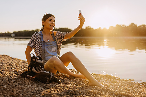 A young Caucasian woman is sitting on the beach with roller skates by her side, taking a picture of herself with her mobile phone.
