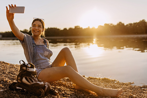 A young Caucasian woman is taking a picture of herself at the beach, with the sunset in the background and roller skates by her side.