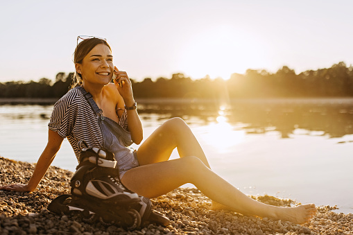 A young Caucasian woman is resting at the beach and having a phone call while smiling wide.