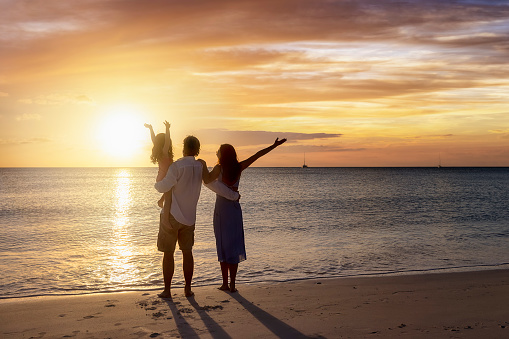 A happy family with outstretched arms on vacation stands on a beach during sunset time