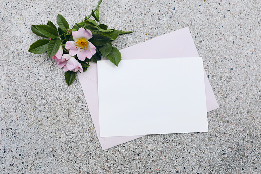 Summer wedding stationery mock-up scene. Horizontal blank greeting card, invitation, pink envelope and blooming dog rose flowers on textured terrazzo background, flat lay, top view. No people.