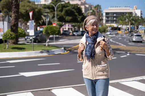 Happy caucasian senior woman crossing the street walking in a sunny city centre expressing positivity, good mood, holding backpack.