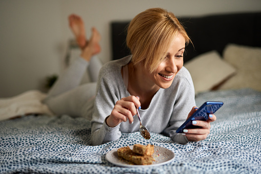 Young women lying on bed, eating cake and using mobile phone