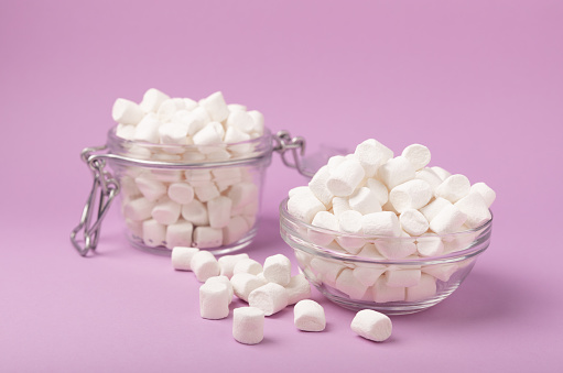 Marshmallows in a glass bowl and a jar on a lilac background.Sweets and snacks for a snack.Chewy candy close-up.Copy space.Place for text.Marshmallows in a glass bowl and a jar on a lilac background.Sweets and snacks for a snack.Chewy candy close-up.Copy space.Place for text.