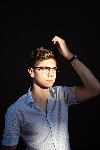 Attractive young man with modern eyeglasses and hairstyle posing on beautiful afternoon sunlight against deep shadow dark background. Handsome guy fixing his haircut.