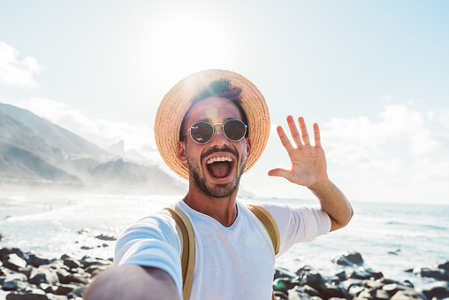 Young man with backpack taking selfie portrait outside - Smiling happy guy enjoying summer holidays at the beach - Millennial showing victory hands symbol to the camera - Youth and journey