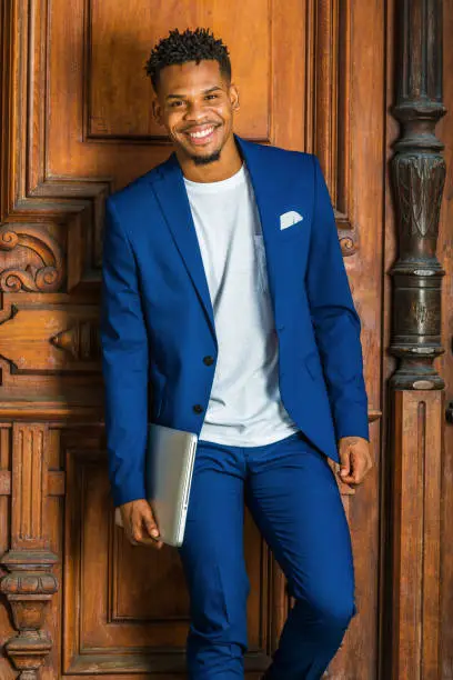African American Businessman working in New York. Wearing blue suit, white T shirt, college student with little goatee, standing by vintage library door on campus, carrying laptop computer, smiling."n