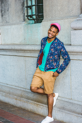 African American Man Casual Street Fashion in New York, wearing blue flower patterned jacket, green V neck T shirt, yellow brown shorts, white sneakers, pink cap, standing by vintage wall, smiling.