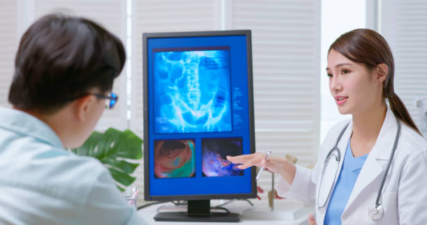 doctor explain colon xray attractive young brunette ponytail female asian doctor wear white coat and stethoscope pen pointing explaining xray film on computer to male patient at clinic - an colon model in background colonoscopy stock pictures, royalty-free photos & images