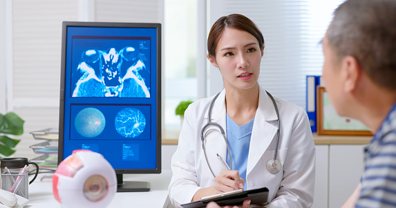 attractive young brunette ponytail female asian doctor wear white coat with stethoscope asks gray hair old sick man his eye condition writing it down at clinic - an eye model and xray film display