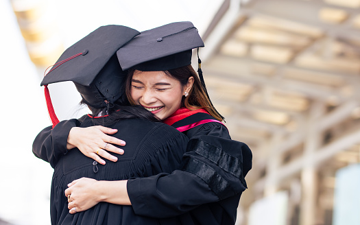 Two asian female students wearing uniform, cap and smiling, hug each other with happiness after graduation. Education Concept