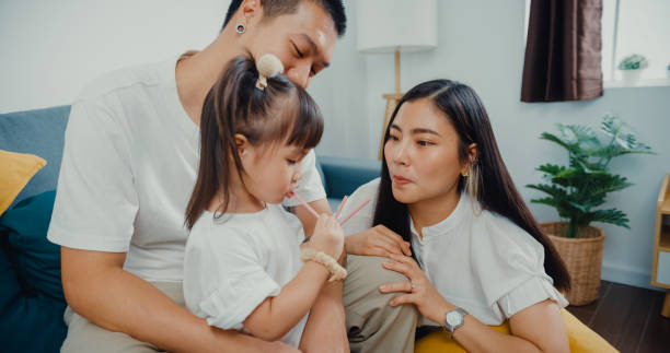 los jóvenes padres asiáticos de la familia juegan con su hijo y comen palo de galletas disfrutan de un feliz momento dorado juntos en la sala de estar de su casa. - eating child cracker asia fotografías e imágenes de stock