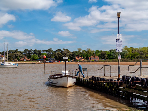The ferryman stepping from the jetty into the motorised ferry boat at Felixstowe Ferry (Old Felixstowe) on a sunny day in springtime. Across the River Deben estuary is the hamlet of Bawdsey. (Suffolk, Eastern England.)