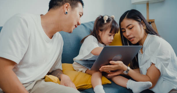 young parent asian family sitting on couch  enjoy with golden time happy moment together with their child use laptop watch cartoon movie in living room at home. - father digital tablet asian ethnicity daughter imagens e fotografias de stock