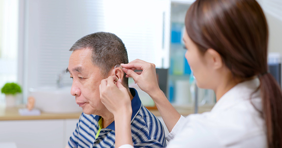 attractive young brunette ponytail female asian doctor wear white coat and stethoscope helps gray hair old man put on hearing aid