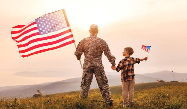 Excited child with american flag holding his father's hand reunited with family Rear view of military man father holding son's hand with american flag and enjoying amazing summer nature view on sunny day, happy male soldier dad reunited with son after US army patriotic man stock pictures, royalty-free photos & images