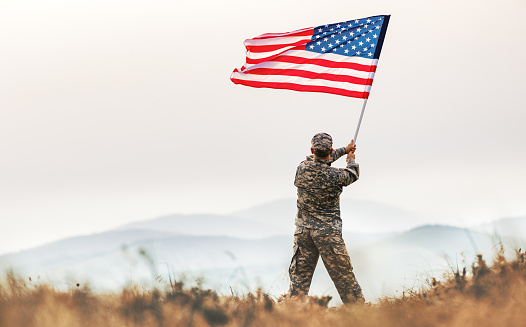 Soldado masculino con el uniforme del ejército estadounidense ondeando la bandera de los Estados Unidos en la cima de una montaña en un claro al atardecer photo
