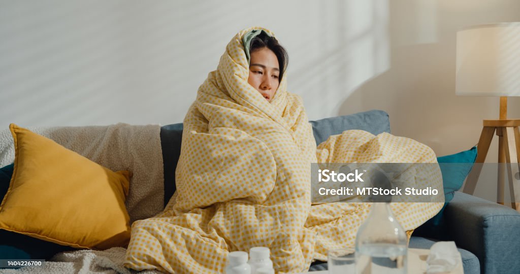 Sick young Asian woman headache fever cough cold sneezing sitting under the blanket on sofa in living room at home. Sick young Asian woman headache fever cough cold sneezing sitting under the blanket on sofa in living room at home. Healthcare concept. Cold And Flu Stock Photo