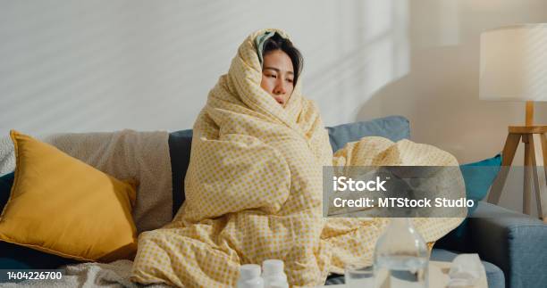 Sick Young Asian Woman Headache Fever Cough Cold Sneezing Sitting Under The Blanket On Sofa In Living Room At Home Stock Photo - Download Image Now