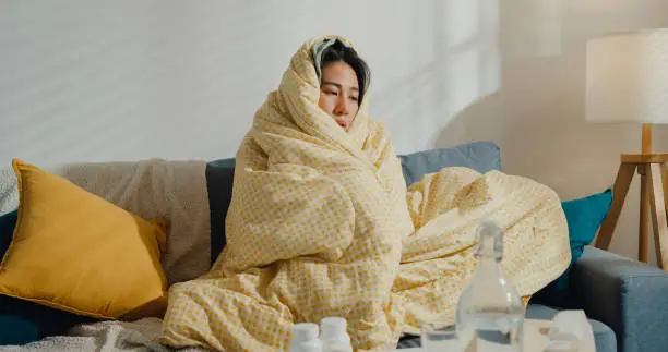 Photo of Sick young Asian woman headache fever cough cold sneezing sitting under the blanket on sofa in living room at home.