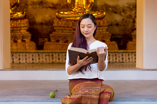 Asian buddhist woman is reading Sanskrit ancient Tripitaka book of Lord Buddha dhamma teaching while sitting in temple to chant and worship inside the monastery