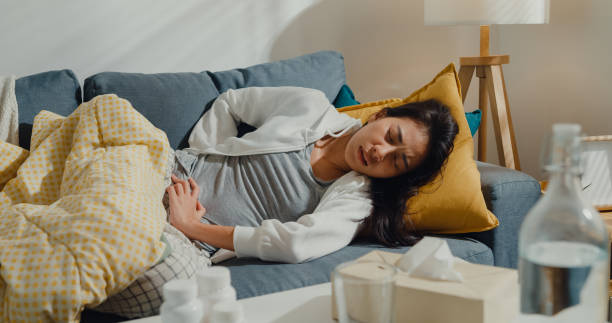 Sick young Asian woman suffering from strong abdominal pain lying down under the blanket on sofa in living room at home. Sick young Asian woman suffering from strong abdominal pain lying down under the blanket on sofa in living room at home. Healthcare concept. Stomach Infection stock pictures, royalty-free photos & images