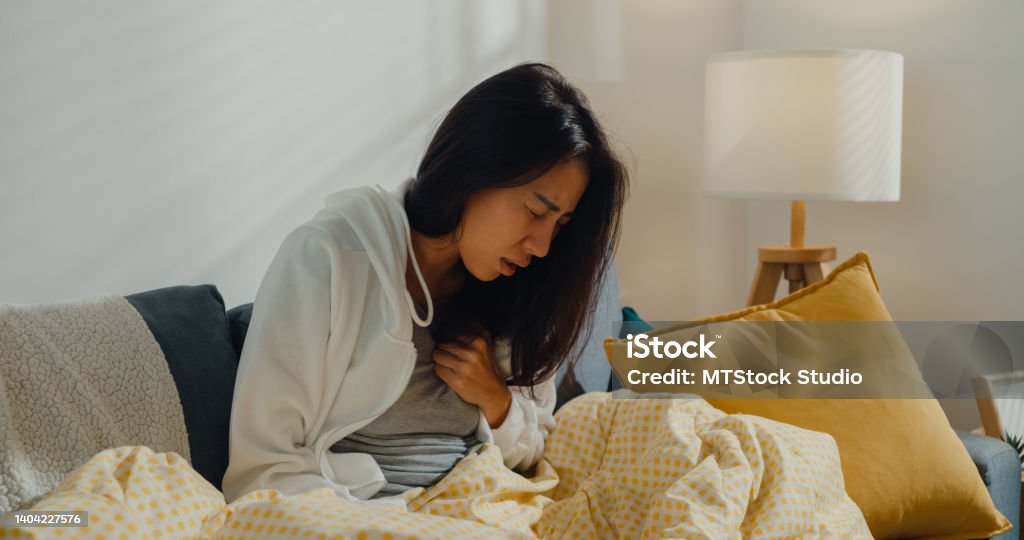Sick young Asian woman having heart attack sitting on sofa in living room at home. Sick young Asian woman having heart attack sitting on sofa in living room at home. Healthcare concept. Tuberculosis Bacterium Stock Photo