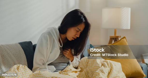 Sick Young Asian Woman Having Heart Attack Sitting On Sofa In Living Room At Home Stock Photo - Download Image Now