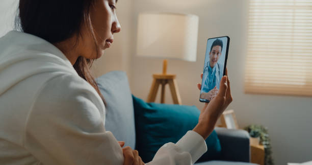 mulher asiática doente usando conversa telefônica com médico sentado no sofá na sala de estar em casa. - cyberspace doctor healthcare and medicine prescription medicine - fotografias e filmes do acervo